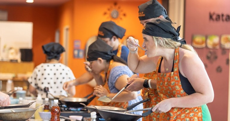 People cooking in a class, wearing aprons and chef hats, tasting food from pans.