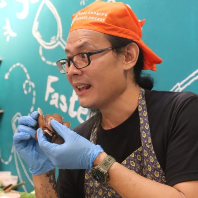 Person with orange bandana and gloves looking upward, standing beside a teal wall with doodles.
